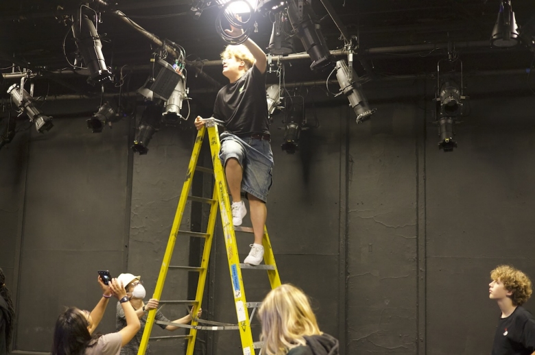 Students watch while an instructor stands on a yellow ladder adjusting stage lights in a studio space 